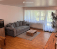 Bright living room with wooden flooring, grey sectional sofa, coffee table, and large windows with white curtains.