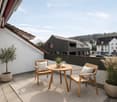 A modern balcony with a small round table, two wooden chairs, and potted plants, overlooking a town with houses and greenery.
