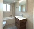 Modern bathroom with a bathtub, toilet, sink, and mirror. Features a wooden vanity cabinet, sleek fixtures, and large windows providing natural light.