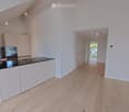 Bright, open-plan space with light wooden flooring, white walls, and a modern kitchen area featuring light-colored cabinets and a black countertop. The room is empty, with no furniture or personal items visible.