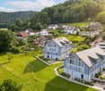 Aerial view of a residential area with multiple houses, surrounded by green fields and trees. The houses have varied architectural styles, with some featuring modern designs and others traditional styles. The area is well-maintained with lawns and gardens