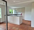 Modern kitchen with white cabinets, a central island, and terracotta tiled flooring. Features a sink with a faucet and large windows allowing natural light.