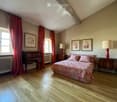 Cozy attic bedroom with wooden floors, large windows, red curtains, a bed with patterned bedding, nightstands with lamps, and framed artwork on the walls.