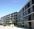 Modern multi-story building with large windows and balconies, surrounded by greenery and parked cars.