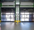 Empty industrial warehouse space with large roll-up doors, metal floor, and control panels visible.