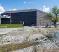Modern building with a sleek, dark facade and large windows, surrounded by a landscaped area with a small stream, trees, and a parking lot.