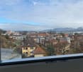View from a balcony showing a town with houses, trees, and distant mountains under a partly cloudy sky.