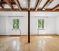 Empty room with wooden herringbone flooring, white walls, exposed wooden beams, and two windows with white frames.