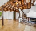 Spacious room with exposed wooden beams, wooden flooring, a brick fireplace, and a staircase leading to an upper level.