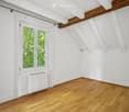 Empty attic room with wooden flooring, white walls, exposed wooden beams, and a large window showing greenery outside.