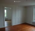 Empty room with wooden flooring, white walls, and large windows. The room is unfurnished, with visible radiators and a doorway leading to another room.