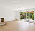 Bright, unfurnished living room with white walls, wooden flooring, fireplace, large sliding glass doors opening to a garden, radiators on walls.