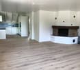 Open-plan living space with light wooden flooring, white walls, and a brick fireplace with a wood stack nearby. The kitchen area features white cabinets and stainless steel appliances.