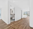 Bright, minimalist hallway with wooden flooring, white walls, and recessed lighting. Features a modern bathroom with a floating sink, toilet, and built-in cabinetry, accessible through an open doorway.