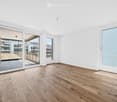 Empty room with light wooden flooring, white walls, large sliding glass doors leading to a balcony, and a view of a modern building complex.