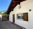 A single-story house with white walls, a wooden door, and a window with wooden shutters. The entrance features a small covered area with a decorative sun-shaped design above the door.