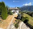 A two-story white house with a gray roof, situated on a hillside with a scenic mountain view in the background. The house features a wooden front door, multiple windows, and a small balcony on the upper level. The surrounding area includes a well-maintain