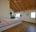 Bright bedroom with wooden floors and slanted wooden ceiling. Features a single bed with colorful heart-patterned bedding, a small desk with a chair, and a mirror on the wall.