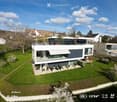 Modern two-story house with a flat roof, large windows, and a spacious lawn. The house features a raised ground floor and is surrounded by greenery and neighboring houses.
