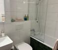 Modern bathroom with white tiles, a bathtub with a glass shower enclosure, a toilet, a sink, and a mirror. Towels and toiletries are neatly arranged on the counter.