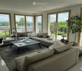 Bright living room with large windows offering a scenic view, featuring a grey sectional sofa, coffee table, armchair, floor lamp, and decorative plant.