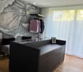 Modern walk-in closet with dark wooden wardrobe, glass top, and hanging clothes. A small stool and picture frame are visible.
