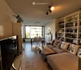 A modern living room with a grey sectional sofa, wooden coffee table, dining area with chairs and table, shelves with books and decorative items, and large windows offering a view of the city.