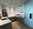 Modern kitchen with white and grey cabinets, a white countertop, and a tiled floor. Features include a sink, faucet, oven, bookshelf with books and a plant, blender, coffee maker, and knife block.