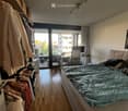 A modern bedroom with a large bed, wardrobe, chair, table, and a balcony view. The room features wooden flooring and a sleek design.
