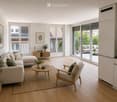 Modern living room with light wood flooring, large windows, and sliding glass doors leading to a balcony. Features a beige sectional sofa, armchair, coffee table, side table, and decorative plants.