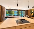 Modern kitchen with a large island featuring an induction cooktop, pendant lights, and a dining area with a table and chairs. A living area with a sofa and ottoman is visible, along with a scenic view of a lake and mountains through large windows.