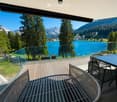 Balcony with wooden decking, glass railing, and outdoor furniture overlooking a lake and mountain landscape.