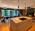 Modern kitchen and dining area with a central island, dining table, chairs, sofa, TV, and decorative vases.