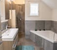 Modern bathroom with a white sink, bathtub, shower, and wooden cabinet. Features a large mirror, decorative tiles, and a small window with a curtain.