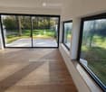 Empty room with wooden flooring and large windows overlooking a garden and pool area.
