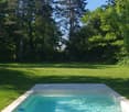 A rectangular swimming pool with clear blue water, surrounded by a tiled deck and lush green grass, set in a serene garden with tall trees.