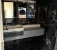 Modern bathroom with black marble walls and floor, white sink, chrome faucet, large mirror, and a washing machine visible in the background.