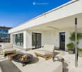 Modern rooftop terrace with white furniture, fire pit, small table with cups, and potted plants, overlooking a scenic view.