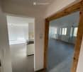 Empty room, wooden door frame, white walls, gray floor, electrical outlet, small kitchen area, radiators, and windows.