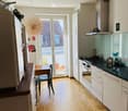 Bright kitchen with white cabinets, black countertop, wooden floor, and a small dining area with a table and chair. Features a modern stove, oven, and a decorative wall piece.