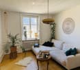 Cozy living room with a white sofa, coffee table, table lamp, and decorative items. Features wooden flooring, large window with white curtains, and several potted plants.