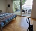 Bright bedroom with wooden flooring, large bed with patterned bedding, a chair, and a table near the sliding glass doors leading to a balcony with outdoor furniture and a scenic view.