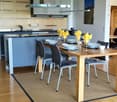 Modern kitchen with a wooden dining table, chairs, and a range hood. The floor is wooden, and there is a beige carpet in the center.