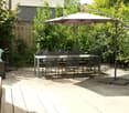 Garden patio with a round table, six chairs, and a large umbrella providing shade. A decorative sculpture sits on a wooden stand to the left. The area is surrounded by lush greenery and a wooden fence.