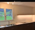 Modern kitchen with grey cabinets, white countertops, a stainless steel sink, and an induction cooktop. Large windows offer a scenic view of a green landscape.