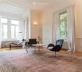 Bright living room with large windows, white curtains, wooden floor, and a patterned rug. Features a black leather sofa, a lounge chair, a small round table with a flower vase, and a floor lamp.