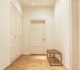 A narrow hallway with light-colored wooden flooring, white walls, and a small wooden bench placed against the right wall. There are several closed doors along the left side.