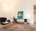 Modern living room with a sleek black chair, a wooden console table holding books and a colorful painting, a floor lamp, and a large area rug on a wooden floor.