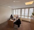 Office space with a wooden desk, black office chair, computer monitor, printer, fan, and a small plant on a cabinet. Large windows provide natural light.