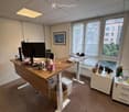 Office space with a standing desk, chair, screen, and various office supplies. There is a small fridge, a white cabinet, and a framed picture on the wall. The room has large windows with a view of the city.